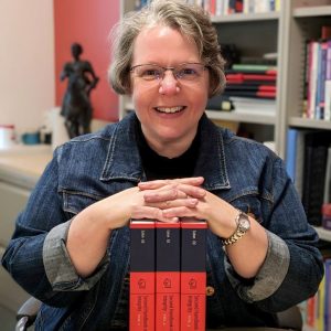 A woman with brown hair and glasses sitting at a table (Sarah Elaine Eaton). She is wearing a denim jacket, a black turtleneck,and a silver-coloured wristwatch on her left wrist. She is leaning on a three-volume set of books with red and navy blue spines. There is white text on the blue portion of the spine and navy blue text on the red portion of the spine. The background is intentionally blurred. There are bookcases on the right-hand side of the background. On the left-hand background, there is a red feature wall, and a beige wall on the far left. There is a table in the left background with a small statue and some other decorations.